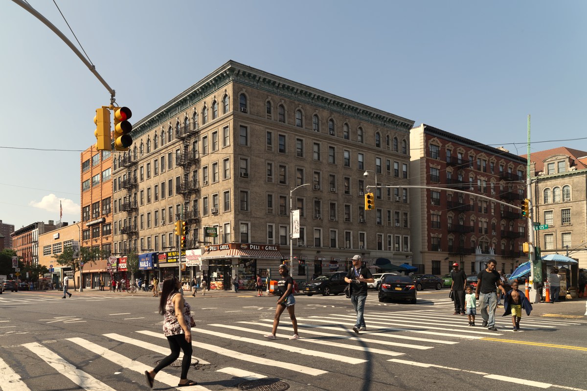 Apartment buildings and shops at a Harlem intersection in New York City, with pedestrians crossing the street